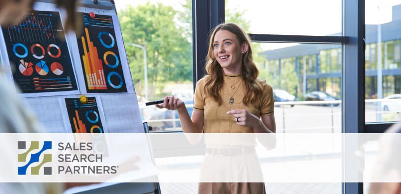 saleswoman with long light hair in a yellow dress points to a board with metics and charts on it in an office meeting room setting